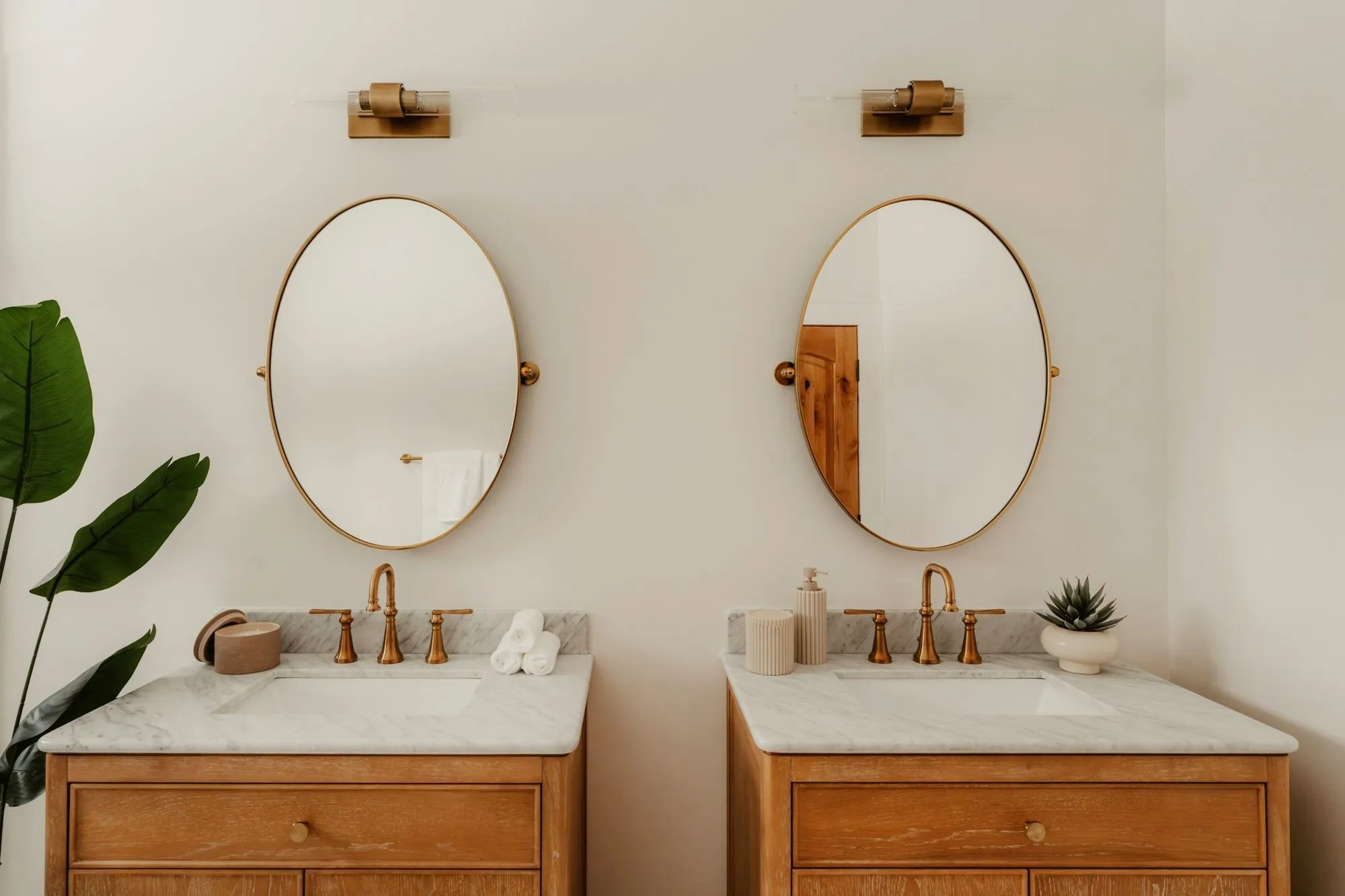 Two oak vanities with marble tops, brass faucets and oval mirrors on a white wall